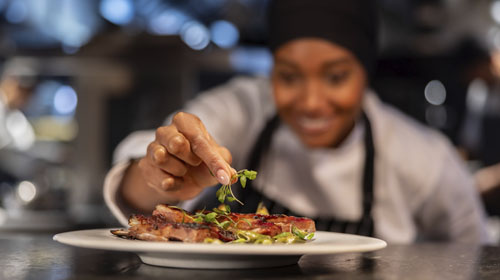 Uma chef de cozinha em um ambiente profissional e moderno finaliza um prato de carne grelhada com ervas frescas, enquanto sorri ao fundo. A cozinha apresenta tons de cinza e prata dos equipamentos de aço inox, contrastando com o preto do avental e o branco do uniforme e do prato. A iluminação quente valoriza os tons dourados e amarronzados da carne e o verde vivo das ervas, criando uma atmosfera sofisticada e acolhedora. A imagem transmite cuidado, técnica e alta gastronomia.